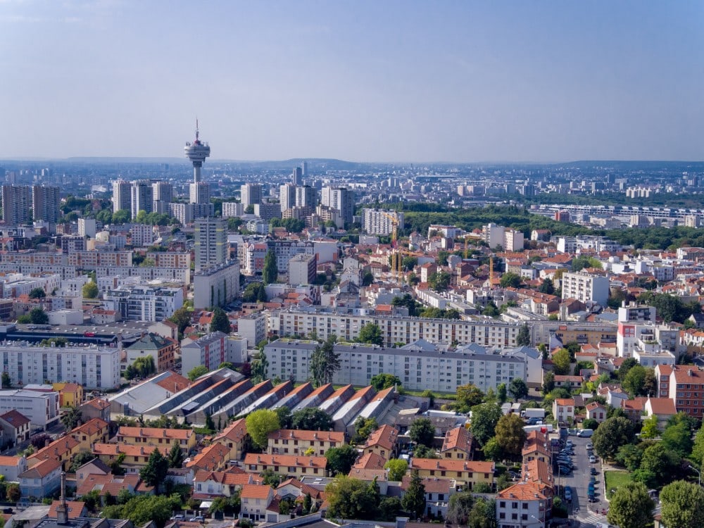 Vue aérienne de Romainville : pavillons aux toits rouges, immeubles collectifs et la tour hertzienne, avec Paris à l’horizon